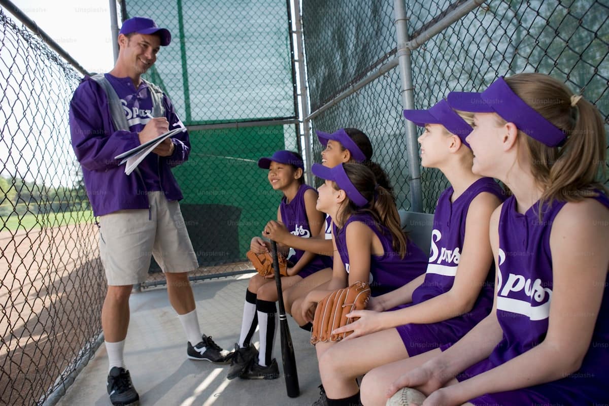 Youth softball coach with clipboard talking to players sitting in the dugout during a game