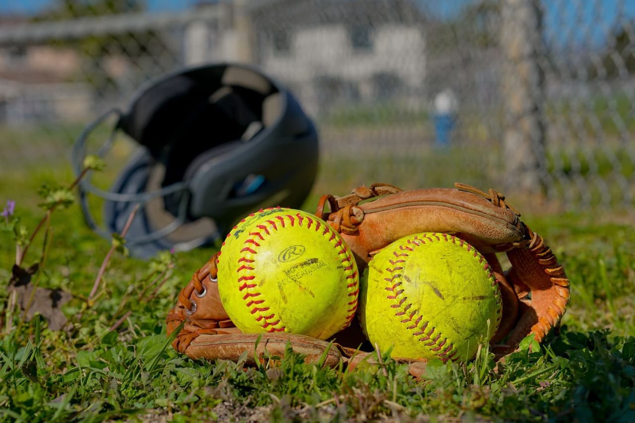 Two yellow fastpitch softballs in a leather glove on the grass, with a batting helmet behind them