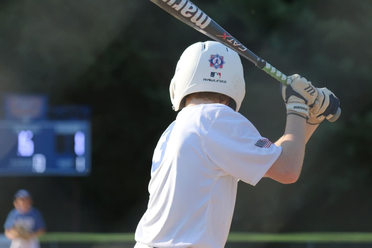 A youth batter in a white helmet loading up at the plate, with the scoreboard blurred behind him