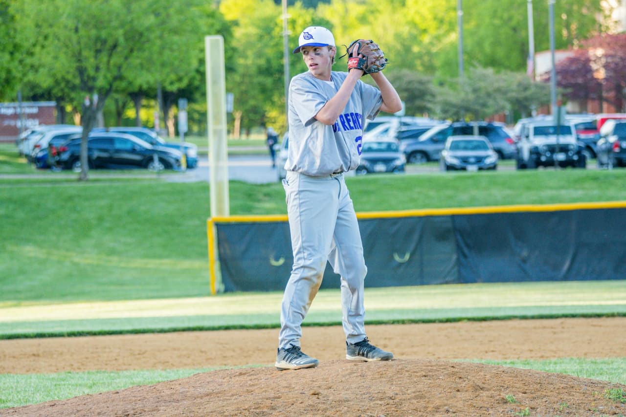 A teenage baseball pitcher in a gray uniform set on the mound with his glove at his chest, looking in before the pitch