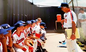 Youth baseball coach arriving early to set up the dugout before players arrive, organizing bat rack and posting lineup card on the fence