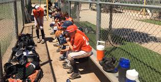 Messy youth baseball dugout with bats on the ground, helmets scattered on the bench, and equipment bags blocking the walkway
