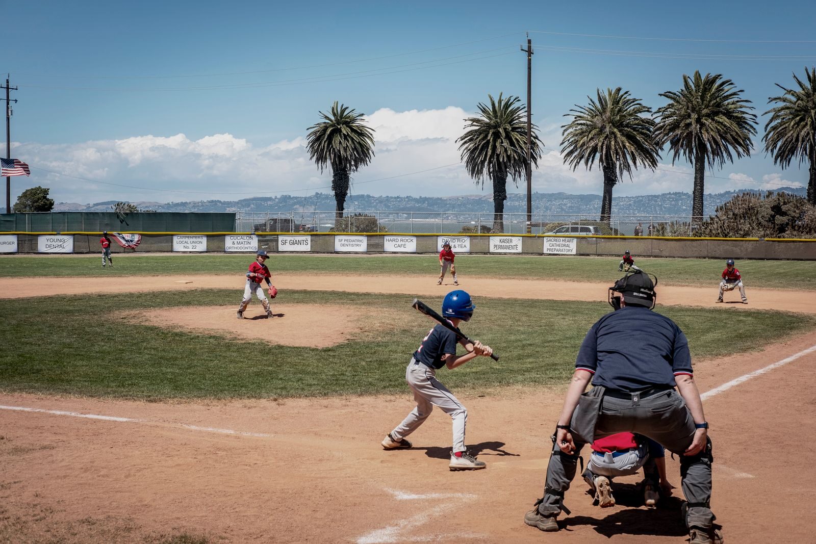Youth baseball game seen from behind home plate, with the batter, catcher and umpire in front and the pitcher and infielders set on a field lined with palm trees