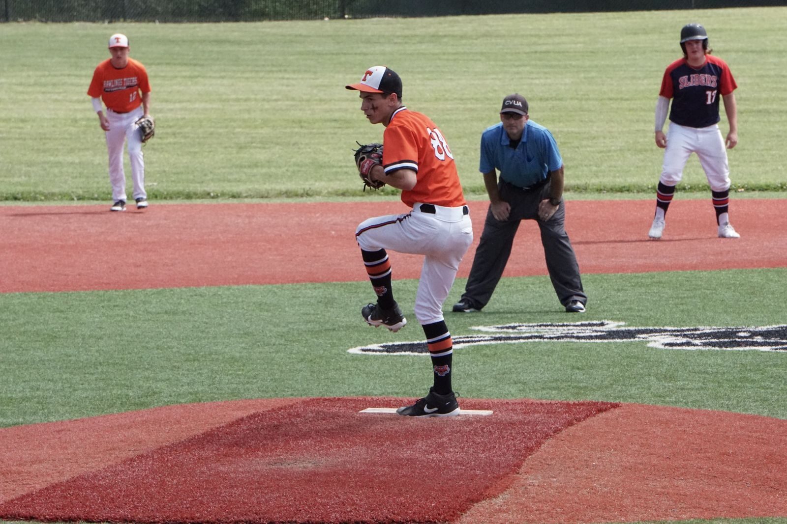 A youth pitcher mid-delivery on the mound during a game, with the umpire and infielders set behind him
